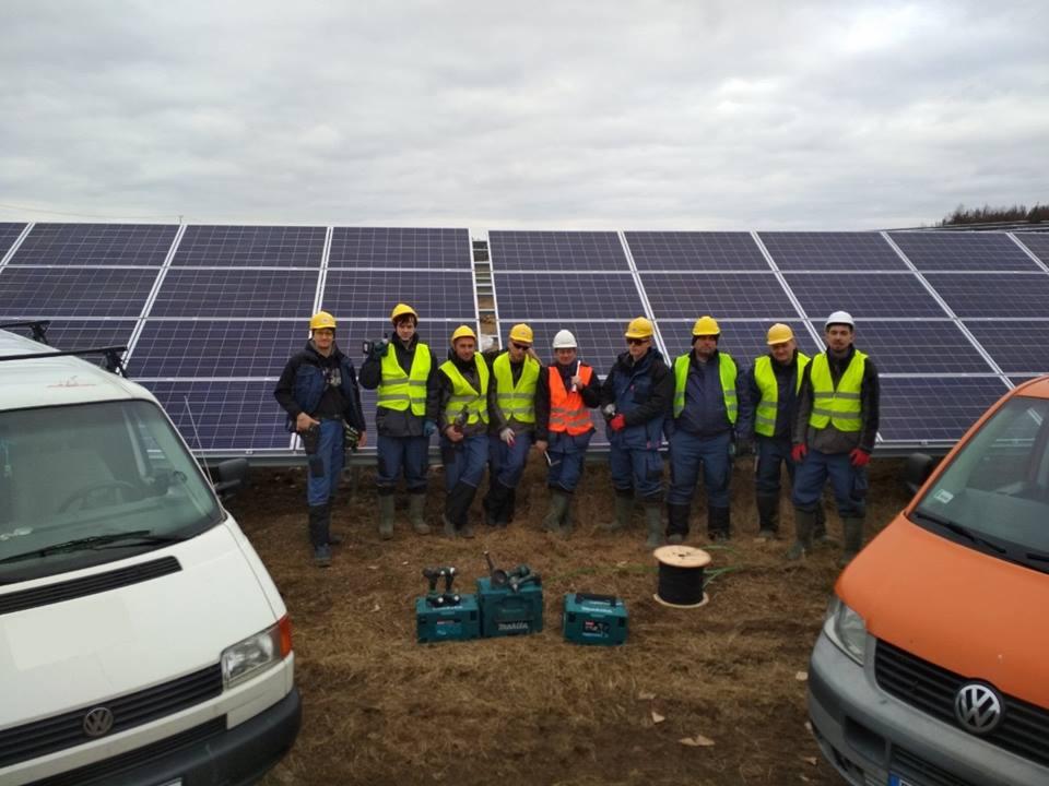 Grupa monterów w kaskach i kamizelkach odblaskowych pozuje na tle zainstalowanych paneli słonecznych, z narzędziami i dwoma samochodami dostawczymi w kadrze.