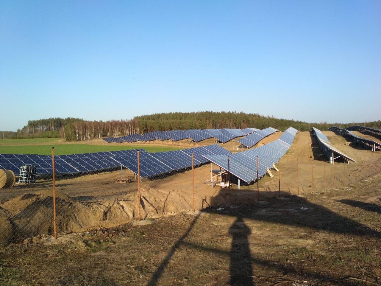Rozległa farma paneli słonecznych na polu, otoczona siatką ogrodzeniową, z lasem w tle i cieniem osoby w dolnej części kadru.