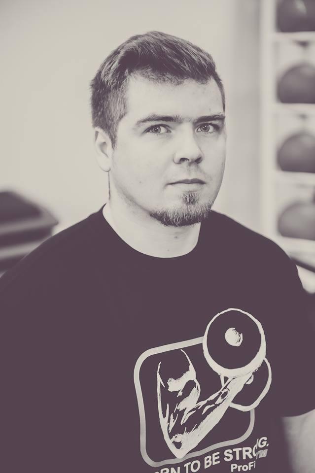 Mężczyzna w czarnej koszulce z nadrukiem bicepsa i ciężarów, stojący na tle jasnej ściany i regału z piłkami.