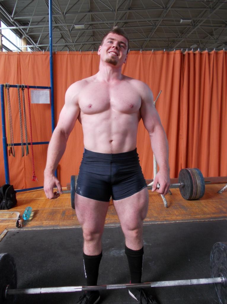 Uśmiechnięty, umięśniony mężczyzna w czarnych spodenkach i skarpetach, prezentujący sylwetkę w siłowni z pomarańczowym tłem i sztangami.