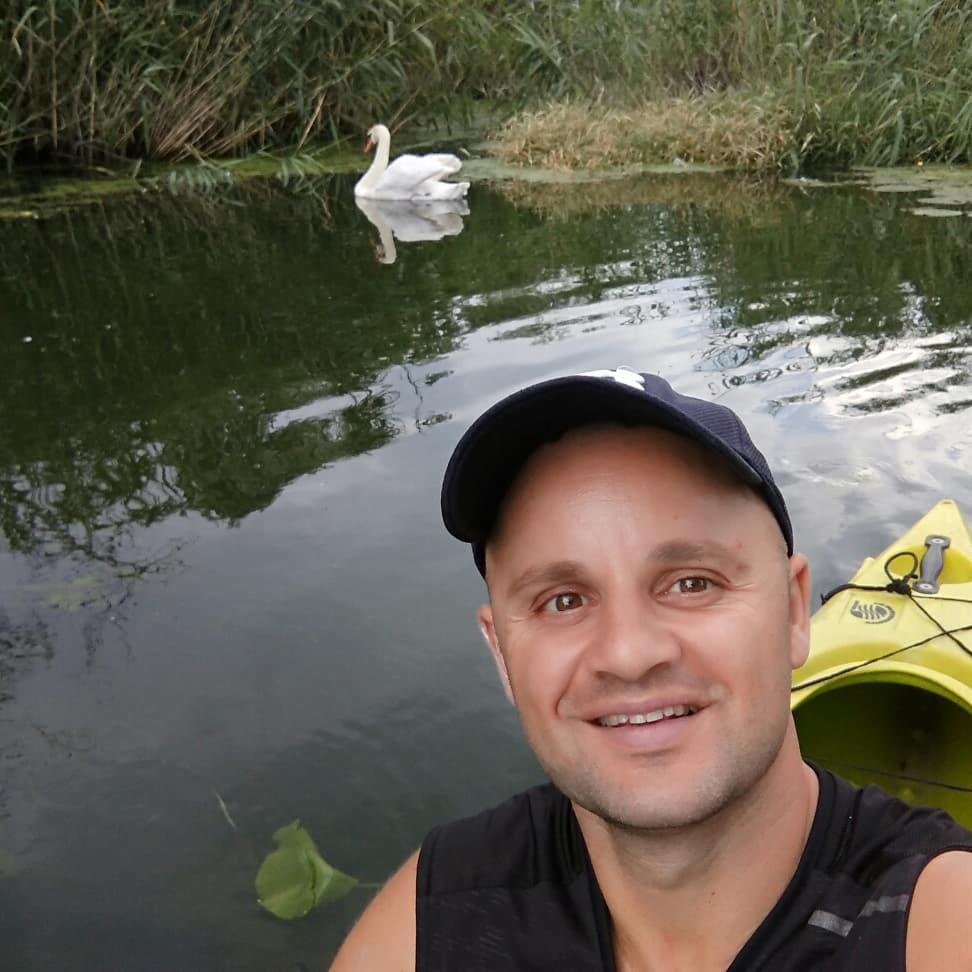 Uśmiechnięty mężczyzna w kajaku, z łabędziem w tle na spokojnej wodzie porośniętej trzciną.