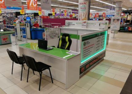 Stanowisko informacyjne z laptopem i krzesłami w supermarkecie, podświetlane zielonym neonem, z widocznymi cenami usług.