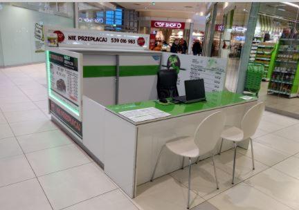 Stoisko informacyjne z hasłem 'Nie przepłacaj' w centrum handlowym, z laptopem, krzesłami i cennikiem.
