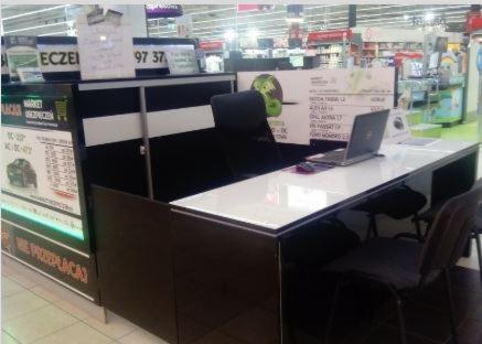 Punkt obsługi klienta z laptopem i informacjami o ubezpieczeniach w supermarkecie, widoczne plakaty reklamowe i tablice informacyjne.