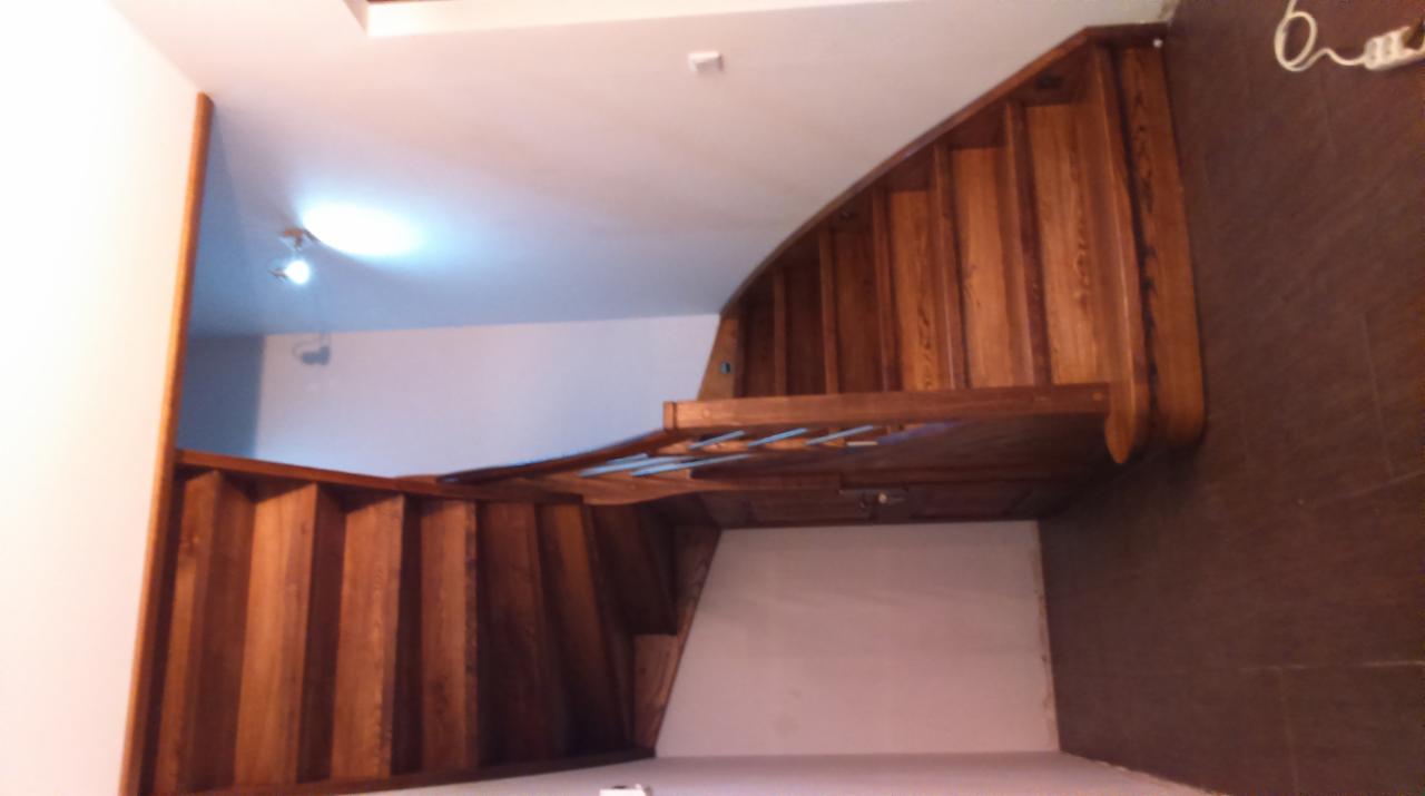 Wewnętrzne schody drewniane z podestem, widok z dołu, ukazujące naturalne usłojenie drewna i zakrzywiony kształt konstrukcji.