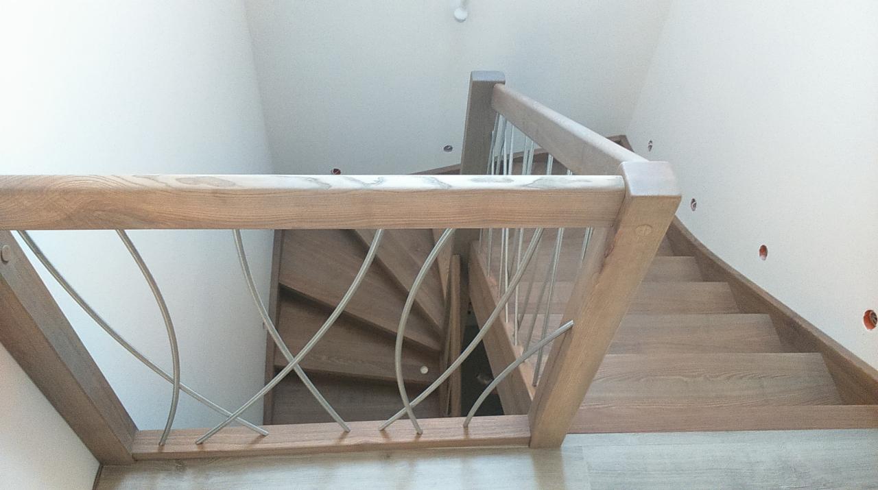 Widok z góry na drewniane schody z balustradą z jasnego drewna i metalowymi prętami o zakrzywionym kształcie, widoczne niedoskonałości na ścianie w postaci otworów.