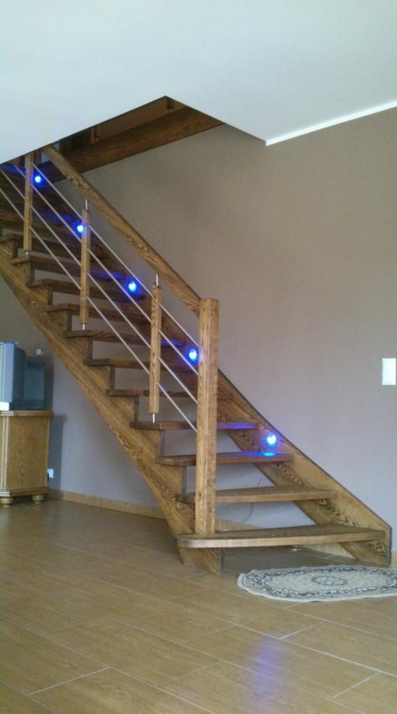 Drewniane schody z metalową balustradą i niebieskim podświetleniem LED, widok z dołu, jasna podłoga z płytek, dywanik u podstawy schodów.