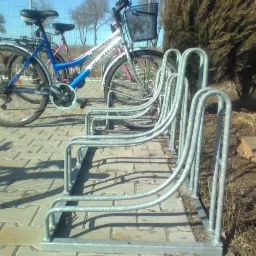 Metalowy stojak rowerowy w kształcie odwróconej litery U, ustawiony na chodniku z kostki, z zaparkowanymi rowerami i krzewem w tle.