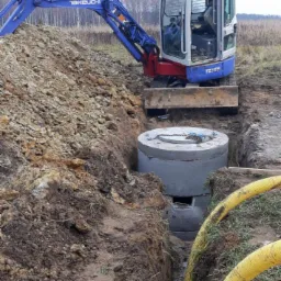 Niebieska minikoparka Takeuchi obok wykopu z betonowymi kręgami, żółte rury w rowie, ziemia i trawa w tle.