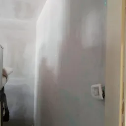 Ściana po nałożeniu pierwszej warstwy szpachli, widoczne niedoskonałości i ślady niebieskiego markera, gniazdko elektryczne częściowo zakryte folią ochronną.