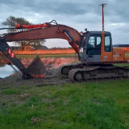 Pomarańczowa koparka Volvo Zaxis 200 LC na brzegu świeżo wykopanego stawu, z widocznymi śladami pracy na gruncie i w wodzie, w tle ogrodzenie i drzewa pod wieczornym niebem.