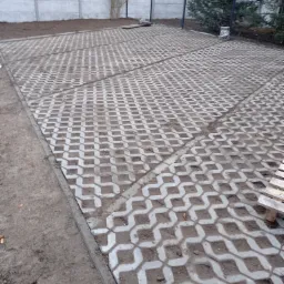 Ułożona szara ażurowa kostka betonowa tworząca wzór na posesji, widoczne wypełnienie ziemią, obrzeża betonowe, w tle ogrodzenie panelowe i betonowe.