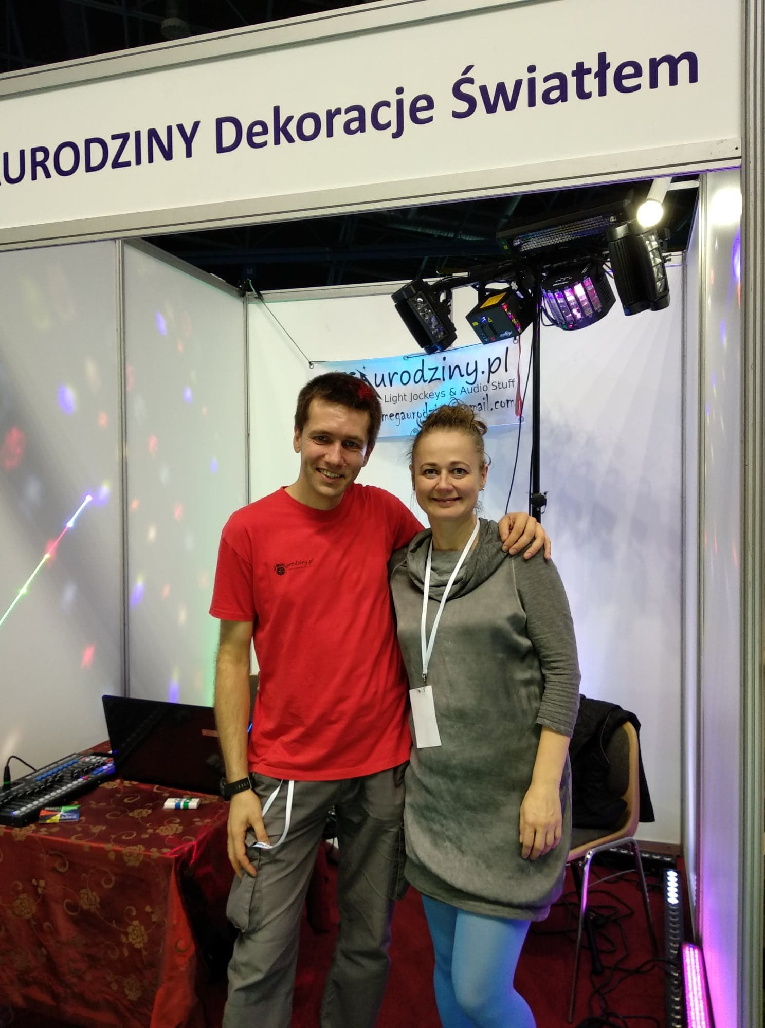 Dwoje uśmiechniętych ludzi na tle stoiska targowego z napisem 'URODZINY Dekoracje Światłem', widoczne oświetlenie sceniczne, laptop i mikser na stole przykrytym wzorzystą tkaniną.