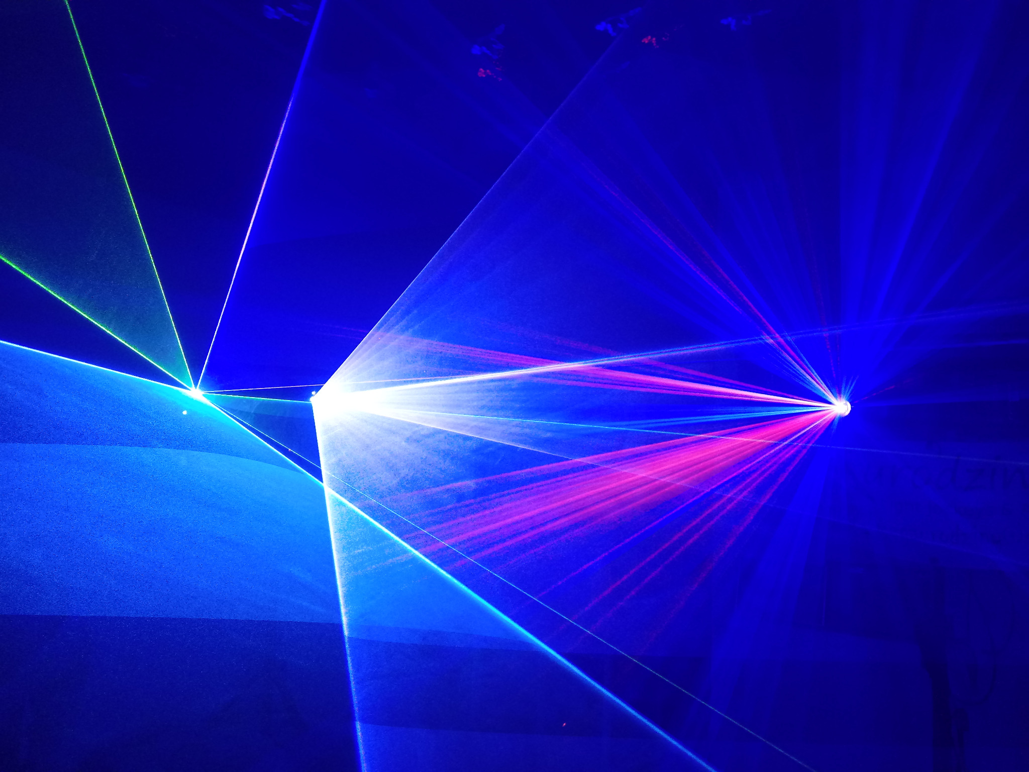 Wiązki różnokolorowych laserów (zielone, niebieskie i różowe) przecinają ciemną przestrzeń, tworząc dynamiczny efekt wizualny.