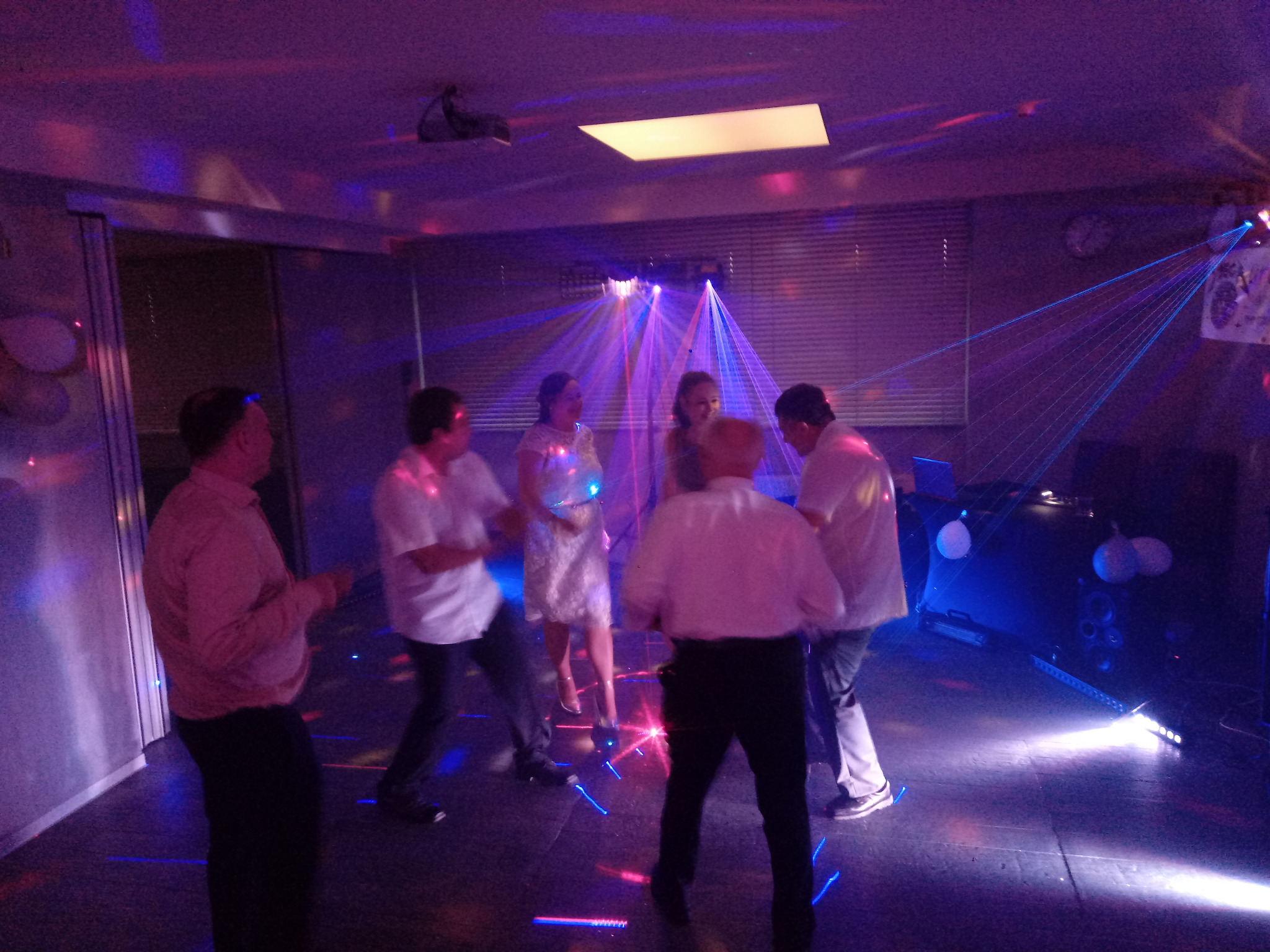 Grupa osób tańczy w pomieszczeniu oświetlonym laserami i kolorowymi światłami; widoczne balony i sprzęt nagłośnieniowy.