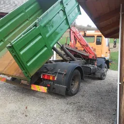 Pomarańczowa ciężarówka z podniesioną, zieloną skrzynią ładunkową i czerwonym dźwigiem hydraulicznym, stojąca na żwirowym podłożu, widok z tyłu i boku.