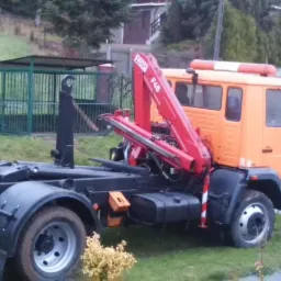 Pomarańczowa ciężarówka z hydraulicznym dźwigiem i platformą do przewozu kontenerów na gruz, stojąca na trawie przed ogrodzeniem.