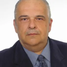 Portret mężczyzny w garniturze i krawacie z subtelnym wzorem, patrzącego prosto w obiektyw na jasnym tle.