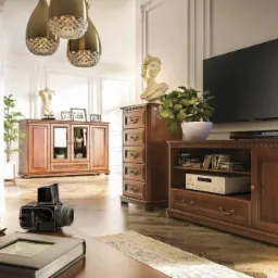 Salon w klasycznym stylu z brązowymi meblami: witryna, komoda z szufladami, szafka RTV pod telewizorem. Na pierwszym planie na stoliku leżą śliwki w miseczce, książka w skórzanej oprawie i aparat...