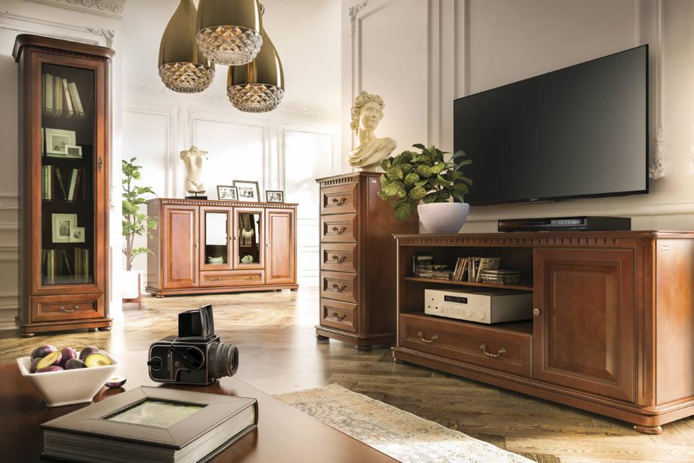 Salon w klasycznym stylu z brązowymi meblami: witryna, komoda z szufladami, szafka RTV pod telewizorem. Na pierwszym planie na stoliku leżą śliwki w miseczce, książka w skórzanej oprawie i aparat...