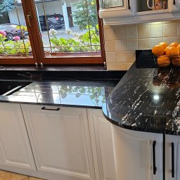 MASTERSTONE - Czarny, błyszczący blat w kuchni z białymi szafkami i czarnym zlewem, widok na okno z kwiatami i odbiciem nieba w blacie.