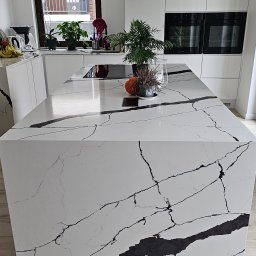 MASTERSTONE - Biała wyspa kuchenna z blatem imitującym marmur calacatta, z czarnymi żyłami, w nowoczesnej kuchni z drewnianą podłogą i białymi szafkami, na blacie rośliny doniczkowe i dekoracje.