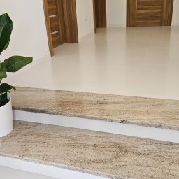 MASTERSTONE - Dwustopniowe schody wewnętrzne wykończone kamieniem w odcieniach beżu i brązu, widoczne z bliska, z rośliną doniczkową po lewej stronie i białymi błyszczącymi płytkami w tle.