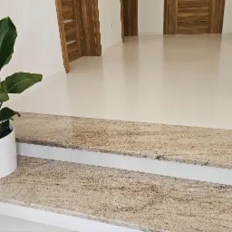 Dwustopniowe schody wewnętrzne wykończone kamieniem w odcieniach beżu i brązu, widoczne z bliska, z rośliną doniczkową po lewej stronie i białymi błyszczącymi płytkami w tle.