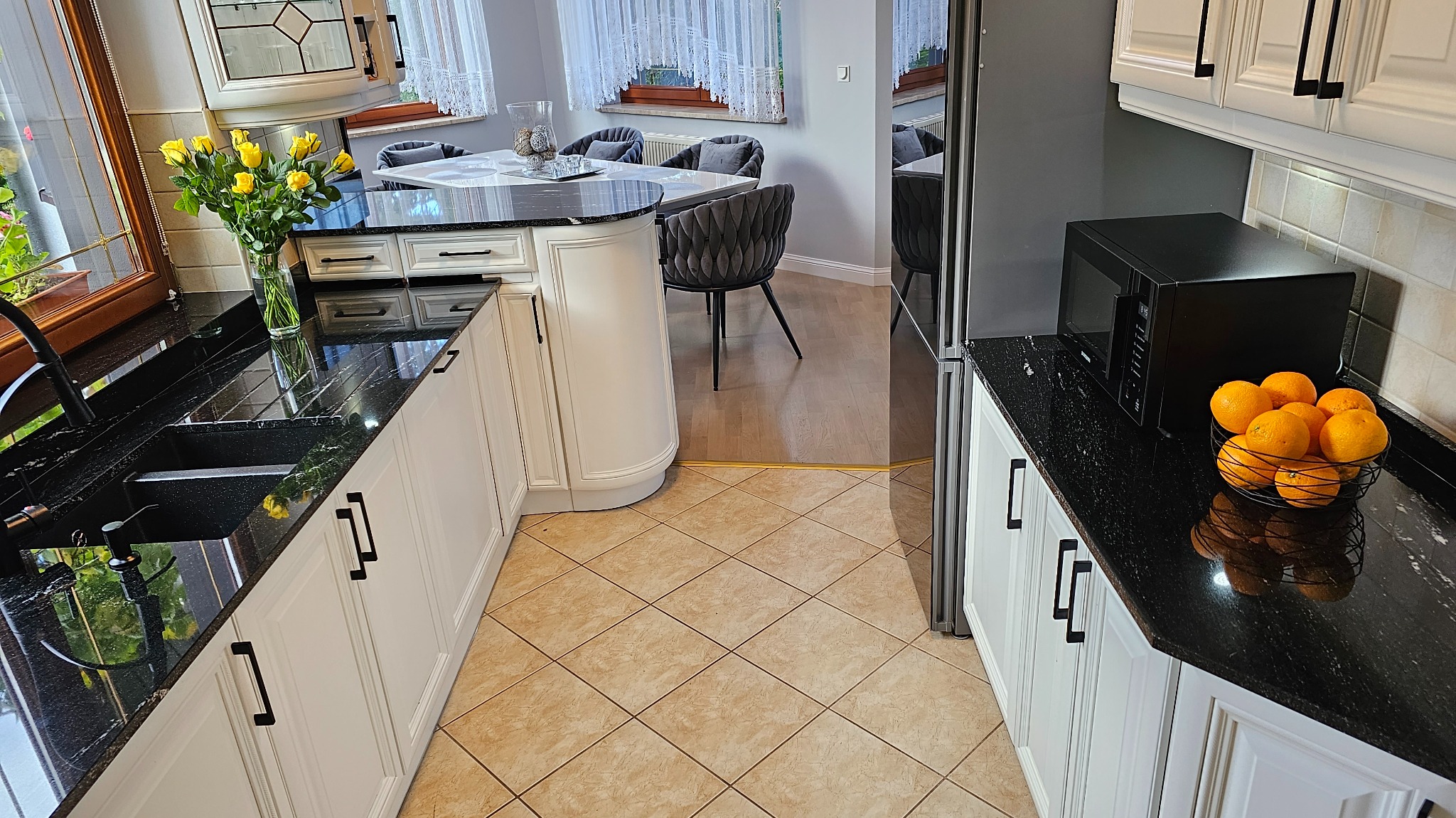 Kuchnia z czarnym blatem kamiennym, białymi szafkami z czarnymi uchwytami, bukietem żółtych róż w wazonie i koszem pomarańczy na blacie, widok na stół z krzesłami w tle.