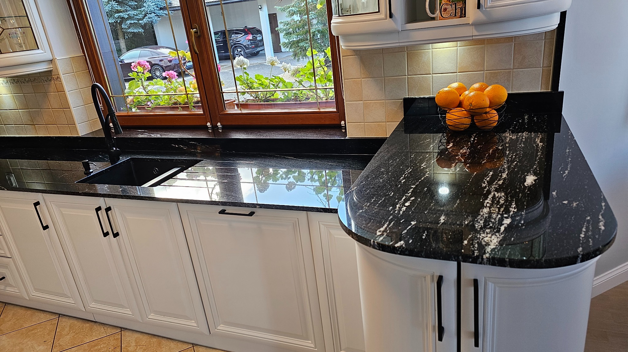 Czarny, błyszczący blat w kuchni z białymi szafkami i czarnym zlewem, widok na okno z kwiatami i odbiciem nieba w blacie.