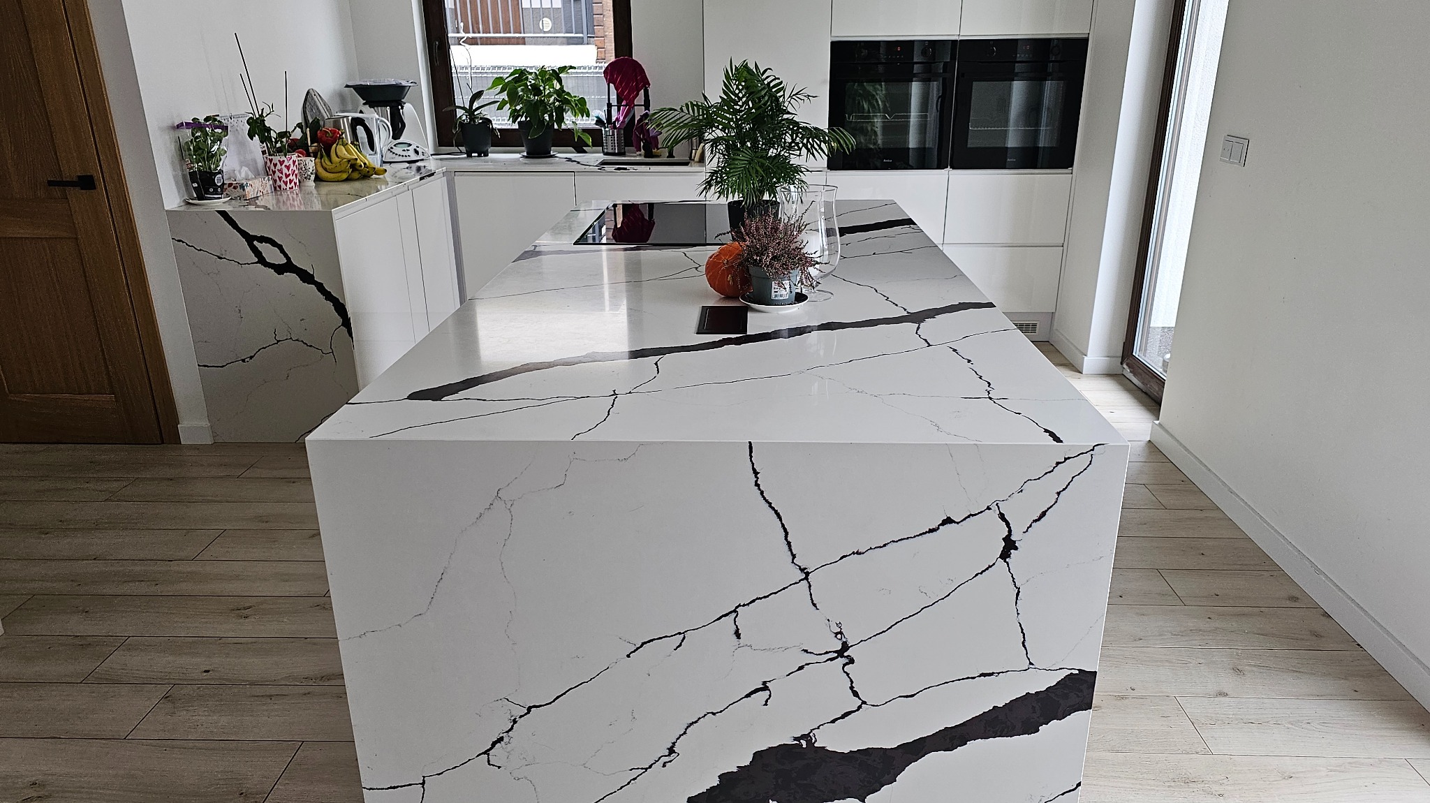 Biała wyspa kuchenna z blatem imitującym marmur calacatta, z czarnymi żyłami, w nowoczesnej kuchni z drewnianą podłogą i białymi szafkami, na blacie rośliny doniczkowe i dekoracje.