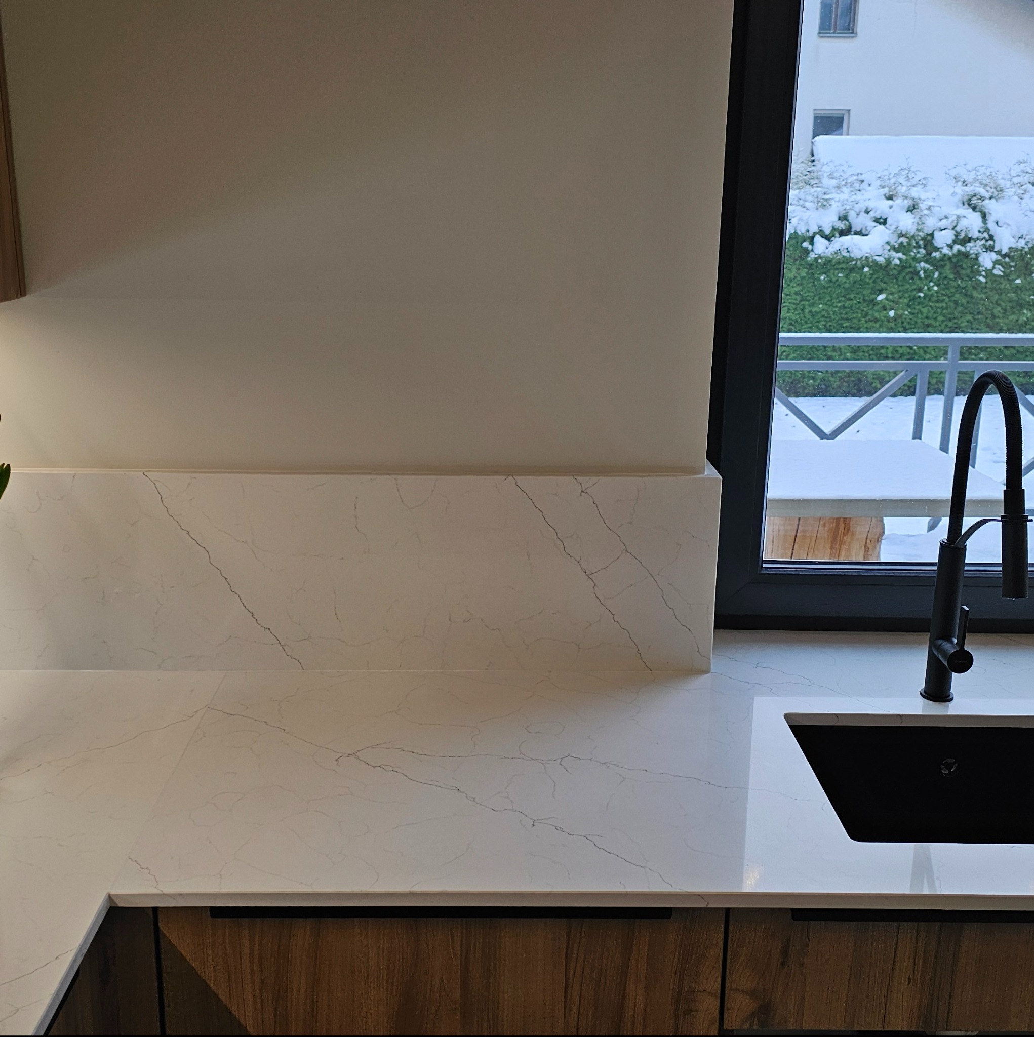 Kuchenny blat z białego konglomeratu z delikatnym marmurkowym wzorem, czarny zlew i bateria na tle okna z widokiem na zimowy krajobraz.