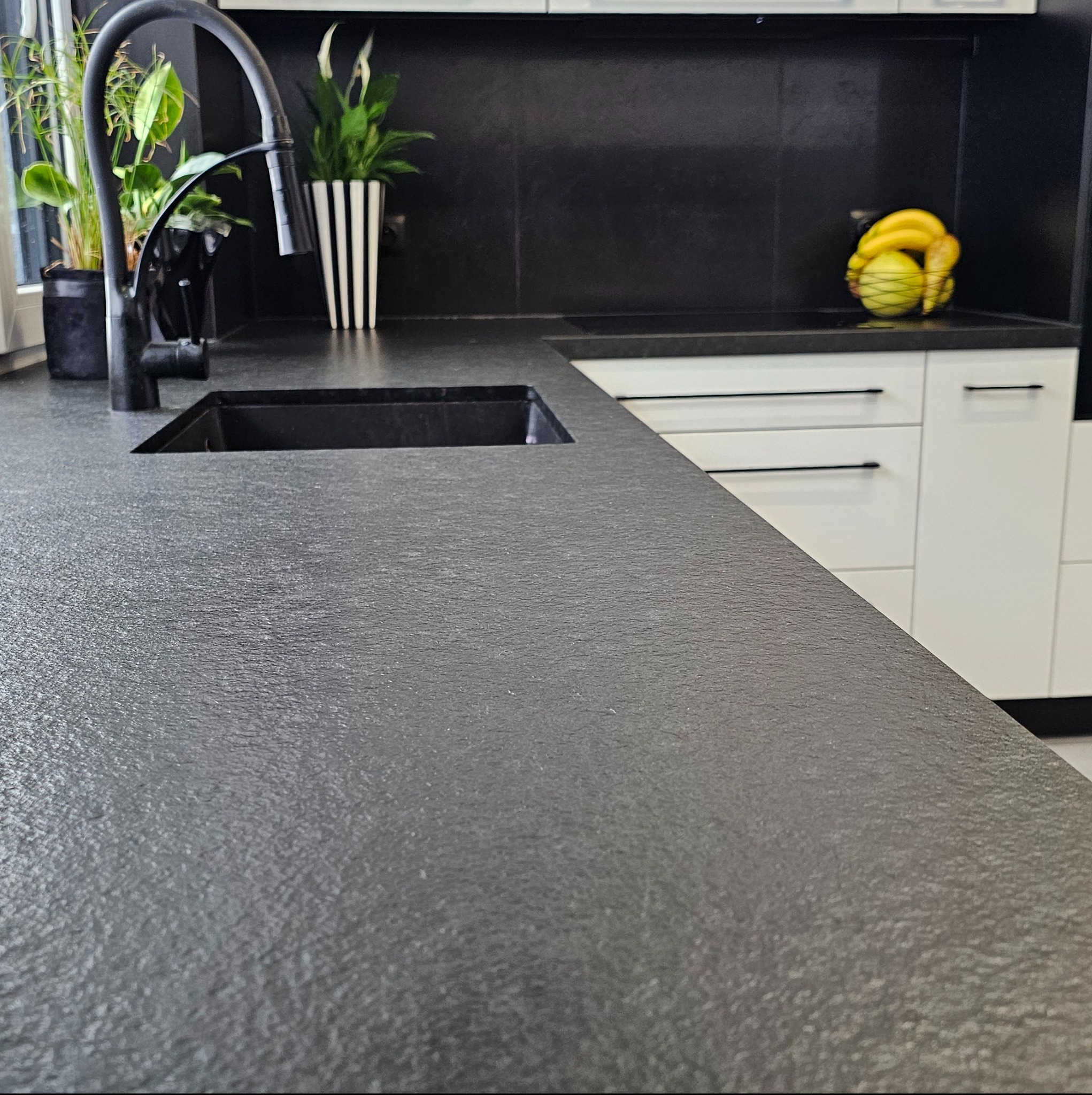 Czarny blat kuchenny z widoczną fakturą kamienia, zintegrowany z czarnym zlewem i baterią, w tle białe szafki i dekoracje roślinne oraz kosz z owocami.