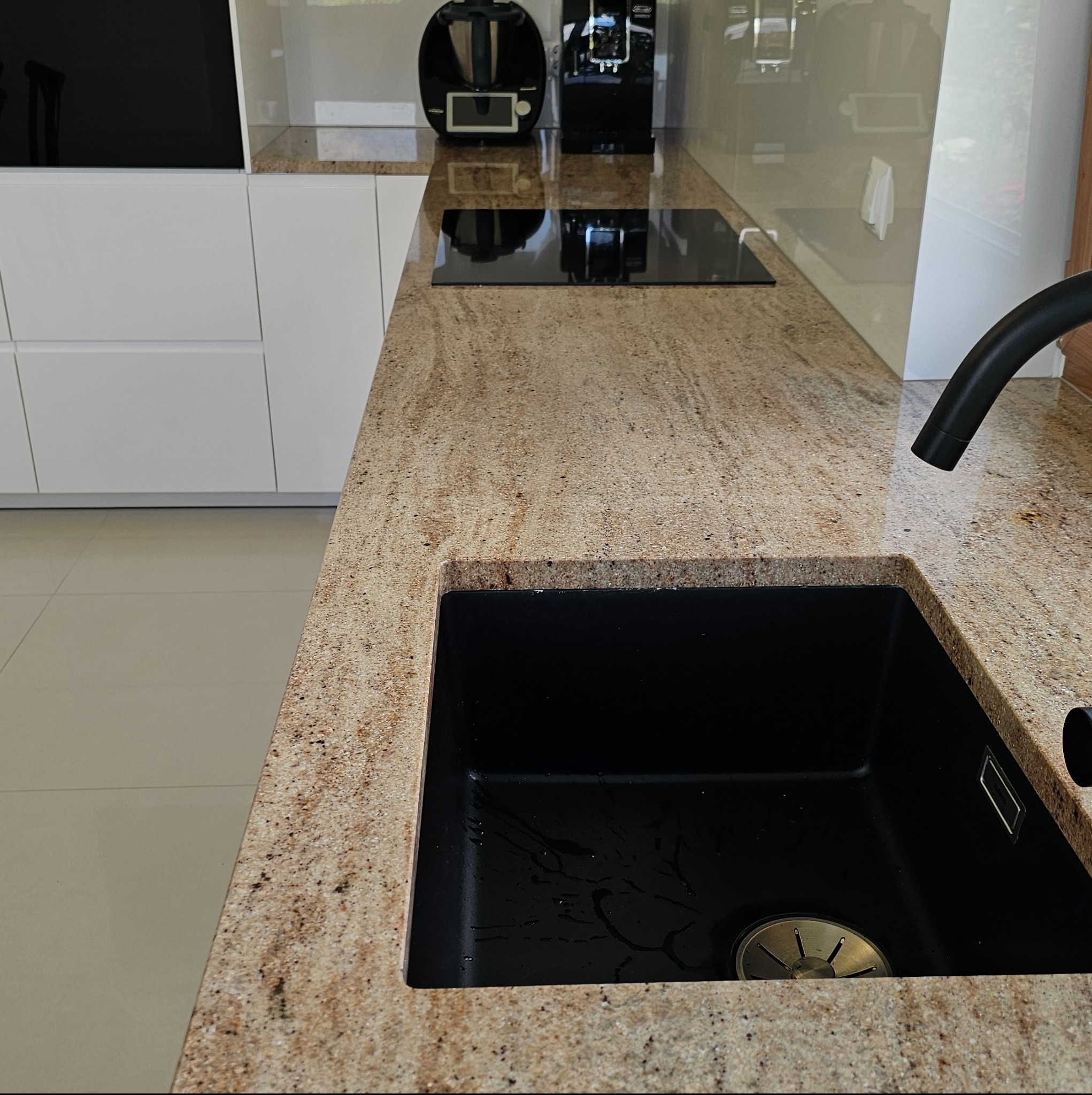 Jasny, ziarnisty blat kuchenny z ciemnym, prostokątnym zlewem i czarną baterią, uzupełniony o ekspres do kawy i Thermomix w tle.