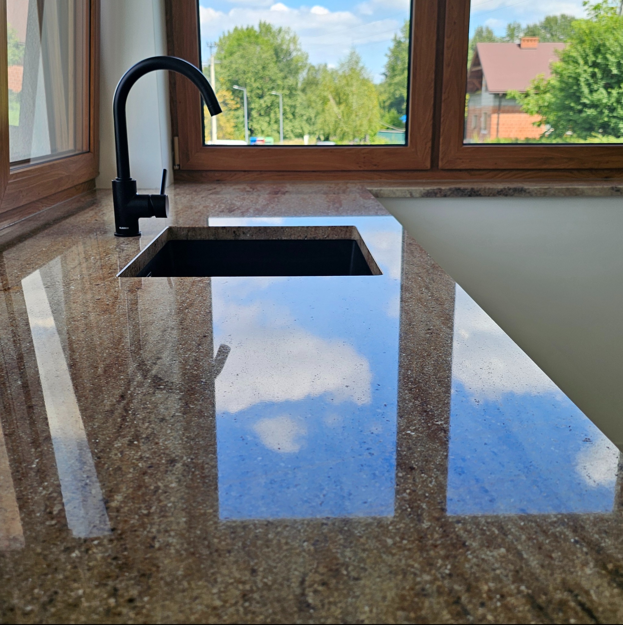 Polerowany blat z brązowego kamienia z czarnym zlewem i baterią, odbijający błękitne niebo, widok przez okno na zewnątrz.