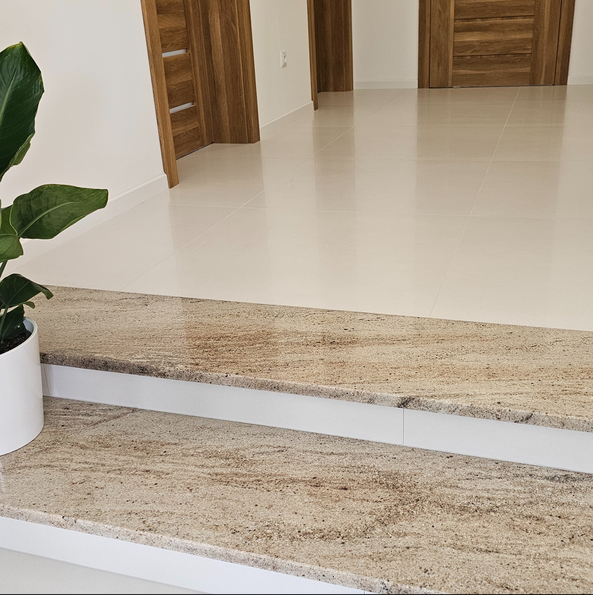 Dwustopniowe schody wewnętrzne wykończone kamieniem w odcieniach beżu i brązu, widoczne z bliska, z rośliną doniczkową po lewej stronie i białymi błyszczącymi płytkami w tle.