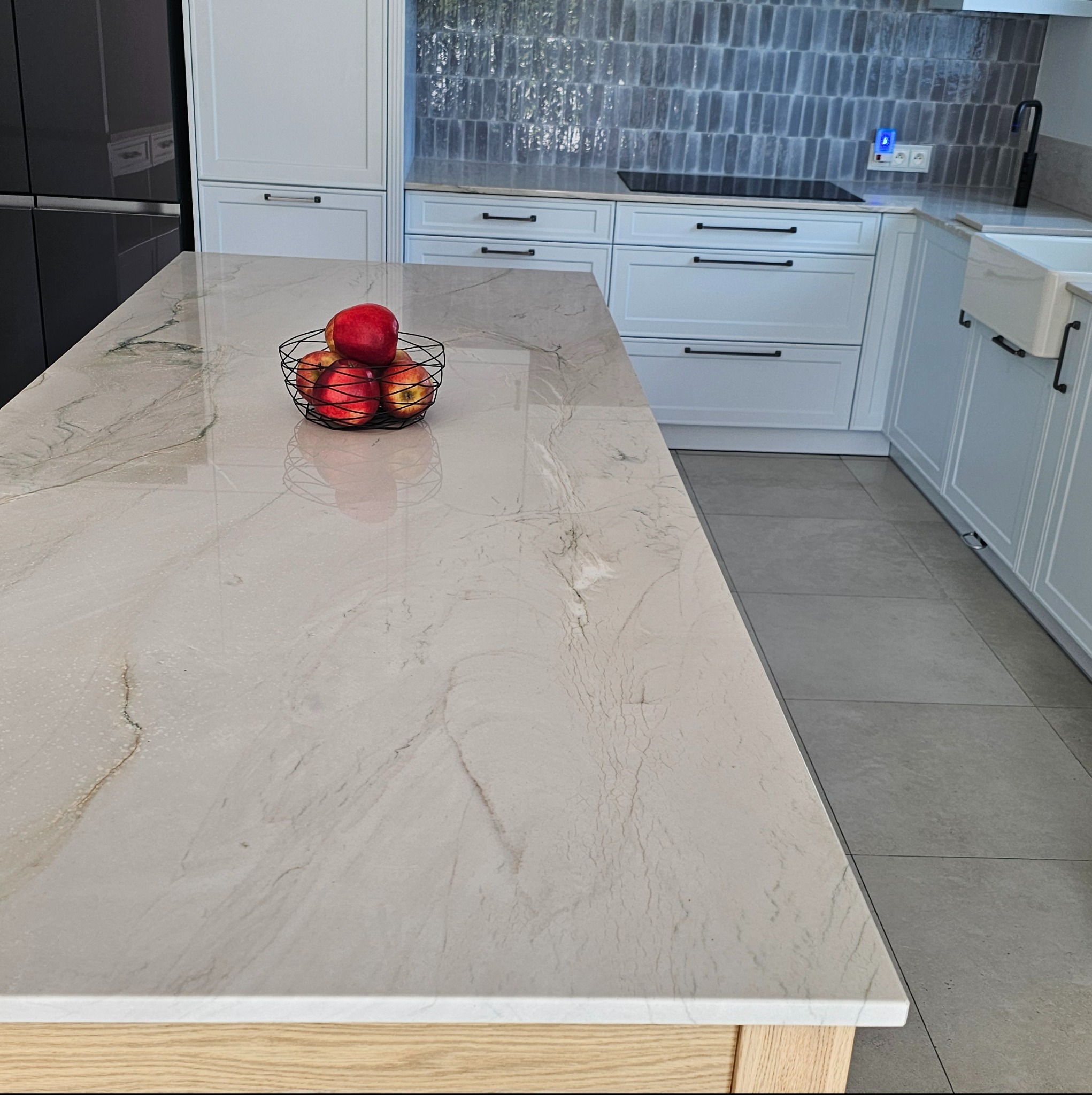 Beżowy blat z delikatnym marmurkowym wzorem w nowoczesnej kuchni z białymi szafkami i koszykiem czerwonych jabłek.