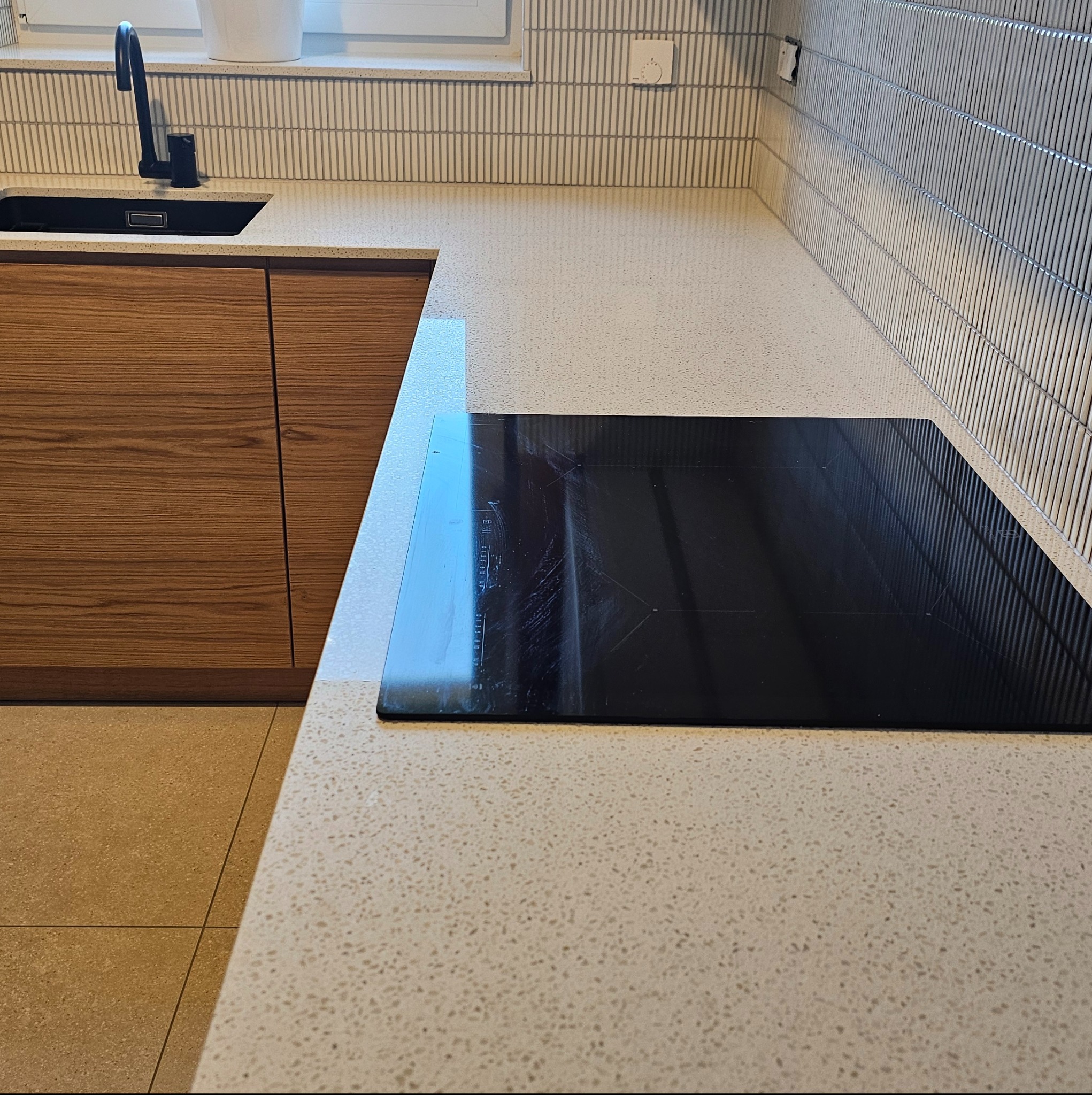 Jasny, nakrapiany blat kuchenny z czarną płytą indukcyjną i zlewozmywakiem, widoczne drewniane szafki i pionowe płytki na ścianie.