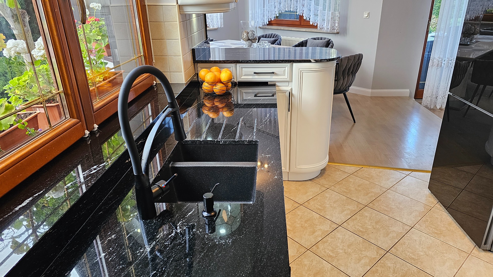 Czarny, błyszczący blat z kamienia z wbudowanym zlewem i baterią w nowoczesnej kuchni, widok z bliska na tle okna z kwiatami i koszyka z pomarańczami.