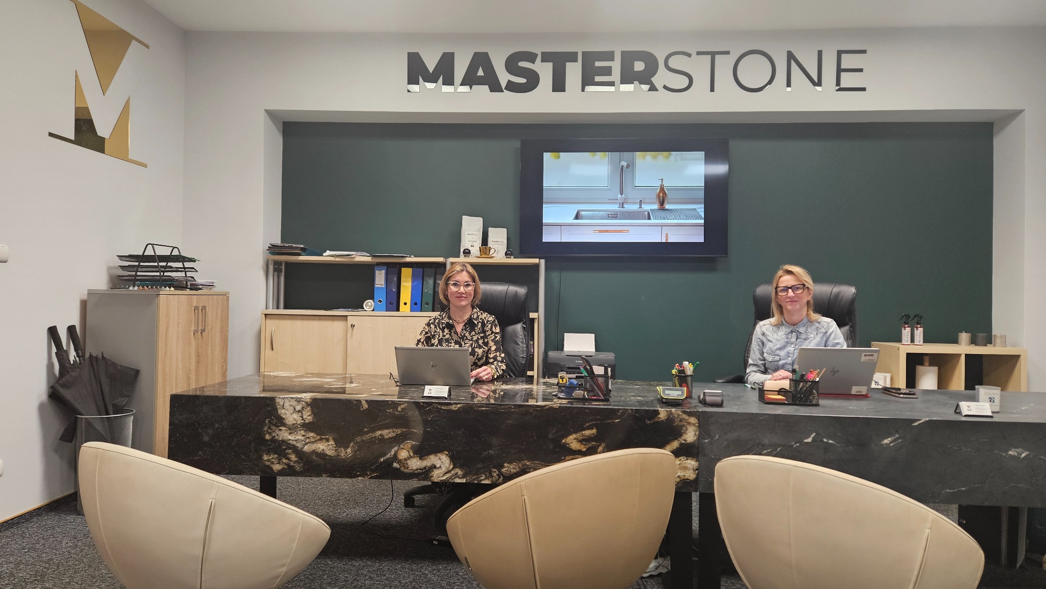 Biuro firmy Masterstone z dwoma pracownicami siedzącymi za biurkami z blatem z ciemnego kamienia z wyraźnym wzorem, na ścianie logo firmy i ekran z wizualizacją blatu kuchennego
