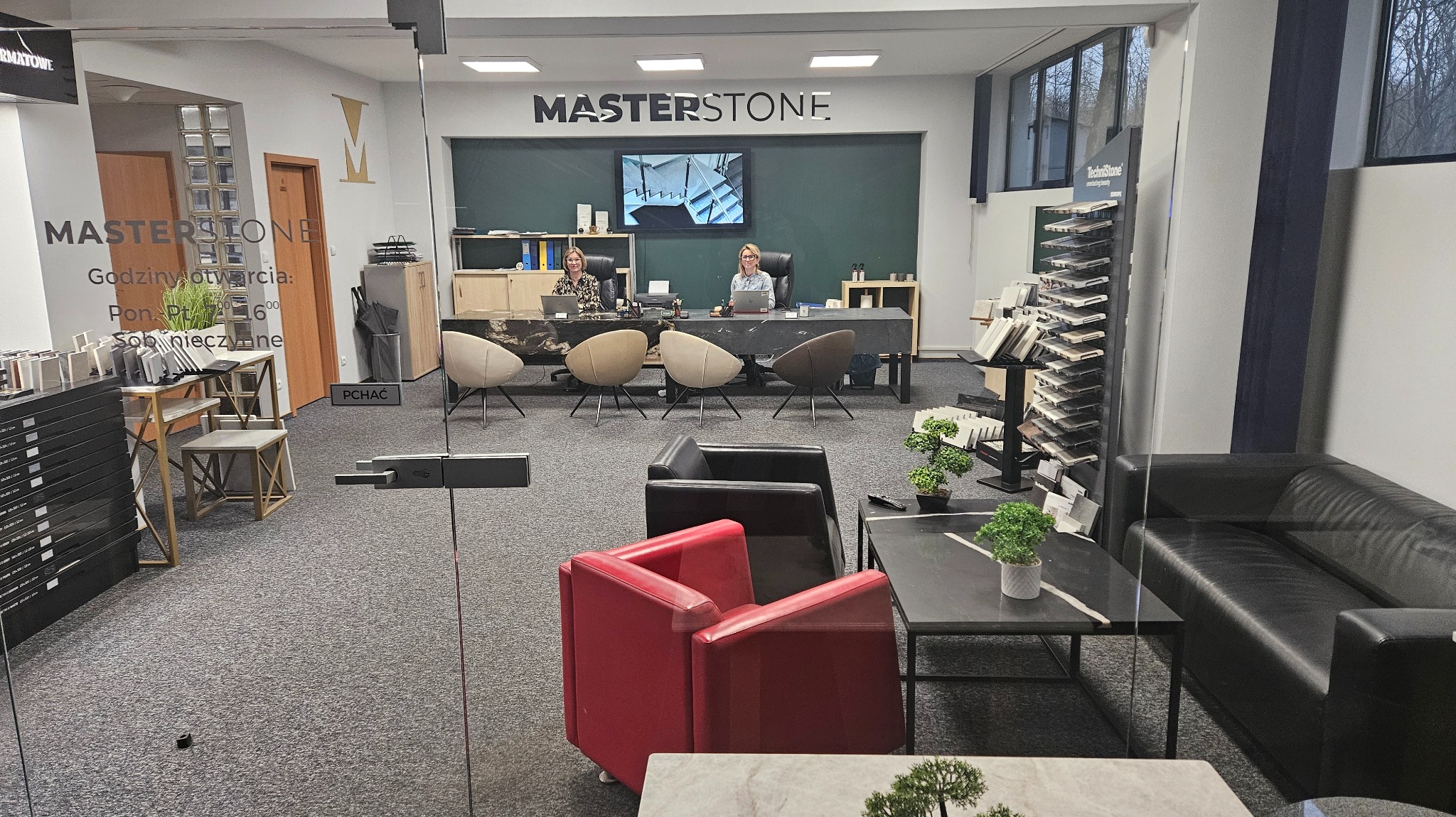 Wnętrze biura firmy Masterstone z ekspozycją blatów kamiennych, widok przez szklane drzwi, dwie kobiety przy biurkach z blatami z kamienia, w tle ekspozytor próbek kamienia.