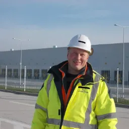 Uśmiechnięty mężczyzna w kasku i żółtej kurtce odblaskowej na tle magazynu z rampami załadunkowymi i błękitnego nieba.