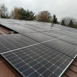 Rząd paneli słonecznych na brązowym dachu w pochmurny dzień, widoczne krople deszczu na powierzchni paneli, w tle drzewa z jesiennym krajobrazem.