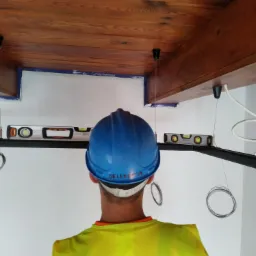 Pracownik w kasku i kamizelce odblaskowej montuje oświetlenie pod drewnianym sufitem, używając poziomicy do precyzyjnego ustawienia. Widoczne kable i uchwyty lamp.