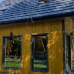 Nowo wybudowany, żółty domek letniskowy z ciemną dachówką, widoczne rusztowanie i klimatyzator na ścianie, otoczony zielenią.