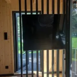 Wnętrze domu z drewnianą boazerią, czarnymi pionowymi listwami dekoracyjnymi, dużym oknem przesuwnym i drabiną opartą o drewniany filar na zewnątrz.