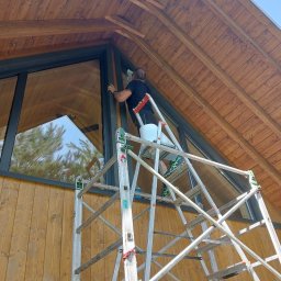 RM SERWIS SPRZATAJACY - Mężczyzna myje okno w domu o drewnianej elewacji stojąc na aluminiowej drabinie z wiadrem, widok z dołu na trójkątny szczyt budynku i odbicie drzewa w szybie.