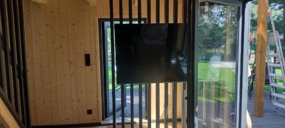 Wnętrze domu z drewnianą boazerią, czarnymi pionowymi listwami dekoracyjnymi, dużym oknem przesuwnym i drabiną opartą o drewniany filar na zewnątrz.