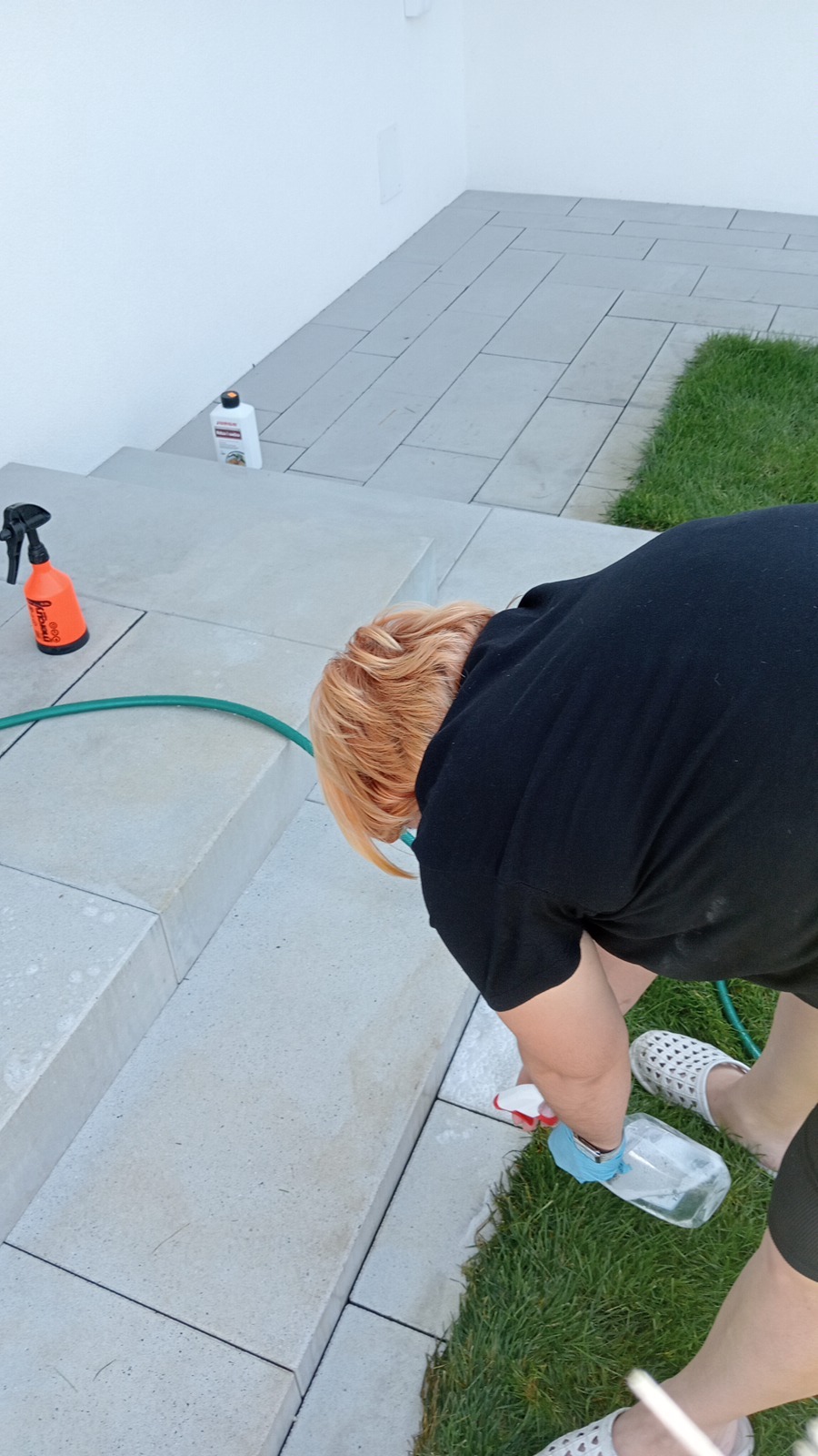 Kobieta spryskuje schody zewnętrzne środkiem czyszczącym, widoczny wąż ogrodowy i butelka preparatu na schodach.