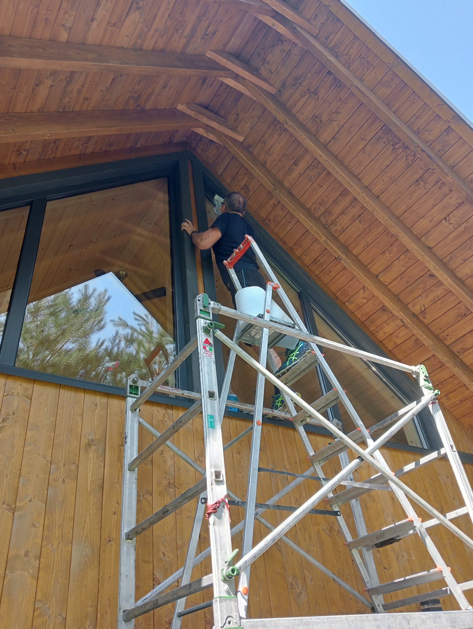 Mężczyzna myje okno w domu o drewnianej elewacji stojąc na aluminiowej drabinie z wiadrem, widok z dołu na trójkątny szczyt budynku i odbicie drzewa w szybie.
